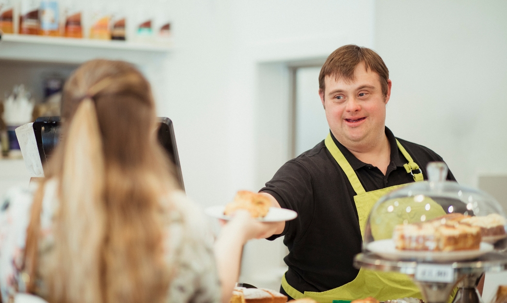 Photograph of a joyful and capable bartender with Down syndrome, wearing a welcoming smile as he proudly serves a delicious croissant on a stylish tray. His positive energy and dedication shine through, embodying inclusivity and diversity in the workplace.