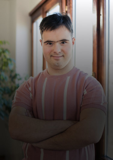 A confident young man with Down syndrome stands with crossed arms, exuding self-assurance and positivity. His expression reflects strength and determination, challenging stereotypes with his empowering presence.